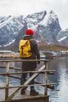 junger Mann mit einem Rucksack, der auf einem hölzernen Pier steht, der Hintergrund der schneebedeckten Berge und des Sees. Platz für Text oder Werbung