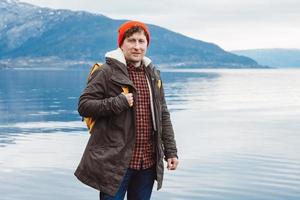 junger Mann mit gelbem Rucksack, der einen roten Hut trägt, der auf einem Felsen vor dem Hintergrund von Berg und See steht. Platz für Ihre Textnachricht oder Werbeinhalte. Reise-Lifestyle-Konzept foto