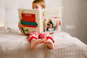 ein Mädchen im ein Weihnachten Schlafanzug ist lesen ein groß Buch auf das Bett, Fokussierung auf ihr Füße foto