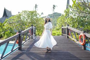Eine glückliche schöne Frau in weißem Kleid, die auf der Holzbrücke über dem Swimmingpool in einem gemütlichen Bungalow mit grünem tropischem Garten auf der Insel Phi Phi, Thailand, genießt und steht