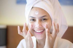 Eine glückliche schöne Frau im weißen Bademantel, die Feuchtigkeitscreme auf das Gesicht im Schlafzimmer, Hautpflege- und Behandlungskonzept aufträgt foto