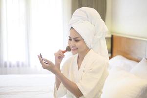Attraktive glückliche Frau im weißen Bademantel trägt natürliches Make-up mit kosmetischer Puderbürste auf, Schönheitskonzept. foto
