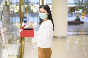 Frau mit Schutzmaske, die eine Geschenkbox im Einkaufszentrum hält, Einkaufen unter Covid-19-Pandemie, Thanksgiving und Weihnachtskonzept. foto