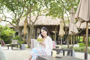 schöne Touristin mit weißer Blume auf ihrem Haar, die während der Sommerferien Kokosnuss trinkt, die auf einem Liegestuhl sitzt