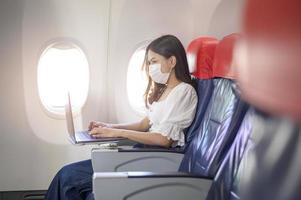 Eine junge Frau mit Gesichtsmaske benutzt einen Laptop an Bord, neue normale Reisen nach dem Konzept der Covid-19-Pandemie foto