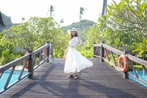 Eine glückliche schöne Frau in weißem Kleid, die auf der Holzbrücke über dem Swimmingpool in einem gemütlichen Bungalow mit grünem tropischem Garten auf der Insel Phi Phi, Thailand, genießt und steht foto