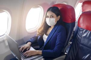 Eine junge Geschäftsfrau, die Gesichtsmaske trägt, verwendet einen Laptop an Bord, neue normale Reisen nach dem Konzept der Covid-19-Pandemie