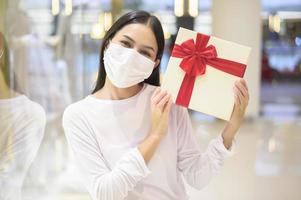 Frau mit Schutzmaske, die eine Geschenkbox im Einkaufszentrum hält, Einkaufen unter Covid-19-Pandemie, Thanksgiving und Weihnachtskonzept. foto
