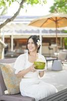 schöne Touristin mit weißer Blume auf ihrem Haar, die während der Sommerferien Kokosnuss trinkt, die auf einem Liegestuhl sitzt