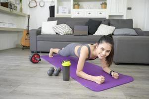 Eine Frau macht Yoga-Plank und sieht sich Online-Trainings-Tutorials auf ihrem Laptop im Wohnzimmer an, Fitness-Workout zu Hause, Gesundheitstechnologiekonzept.