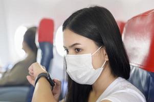 reisende Frau mit Schutzmaske an Bord im Flugzeug mit Smartwatch, Reisen unter der Covid-19-Pandemie, Sicherheitsreisen, Protokoll zur sozialen Distanzierung, neues normales Reisekonzept