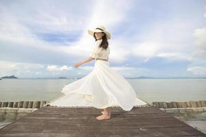 Eine glückliche, schöne Frau im weißen Kleid, die auf der Terrasse der tropischen Insel und dem türkisfarbenen Meer, Sommer- und Urlaubskonzept genießt und sich entspannt