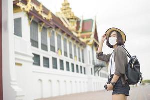 eine junge hübsche asiatische frau trägt eine schutzmaske, die durch berühmte orte in der stadt bangkok reist, neue normale reisen, covid-19 schutz, sichere reisen foto