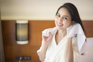 Lächelnde junge Frau mit weißem Bademantel, die sich nach dem Duschen im Schlafzimmer die Haare mit einem Handtuch abwischt