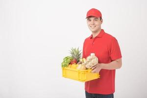 Porträt eines Mannes in roter Uniform hält frische Lebensmittel in einer Plastikbox auf weißem Hintergrund, Hauslieferungskonzept foto