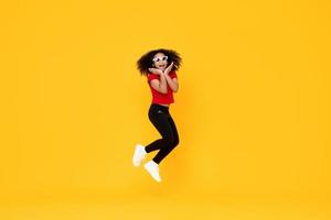 glückliches modisches afroamerikanisches Mädchen, das mit den Händen am Kinn in der Luft springt, isoliert auf gelbem Hintergrund foto