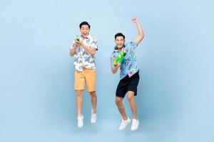 lächeln glückliche asiatische männliche Freunde, die mit Wasserpistolen spielen und auf blauem, isoliertem Hintergrund für das Songkran-Festival in Thailand und Südostasien springen foto