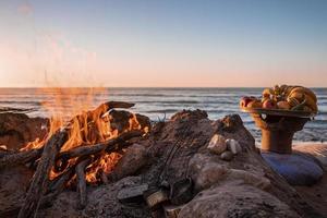 frisches Obst im Teller neben Lagerfeuer mit brennendem Brennholz am Strand am Abend foto