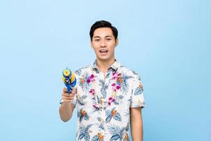 lächelnder gutaussehender asiatischer mann, der mit wasserpistole spielt, isoliert auf blauem hintergrund des studios für das songkran-festival in thailand und südostasien foto