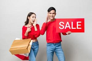 asiatisches Paar mit Taschen und rotem Verkaufsschild in isoliertem grauem Studiohintergrund für chinesisches Neujahrs-Shopping-Konzept