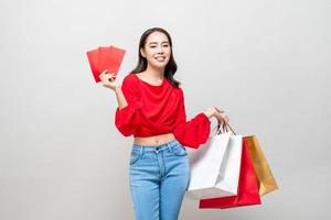 glückliche asiatische frau, die einkaufstaschen und rote umschläge oder ang pao hält, einzeln in hellgrauem studiohintergrund für chinesisches neujahrsverkaufskonzept foto