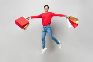 Schöner lächelnder asiatischer Mann, der Einkaufstaschen hält und in isoliertem Studio hellgrauen Hintergrund für chinesische Neujahrsverkaufskonzepte springt
