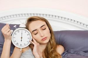 Konzept der verschlafenen Arbeit, Schlaflosigkeit - junges schönes schläfriges Mädchen mit geschlossenen Augen hält eine Uhr und liegt morgens in ihrem Bett im Schlafzimmer