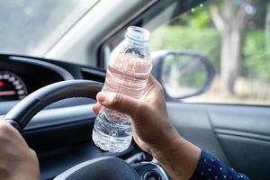 asiatische Fahrerin, die eine Flasche zum Trinken von Wasser hält, während sie ein Auto fährt. Wärmflasche aus Kunststoff verursacht Feuer. foto