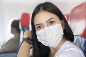 reisende Frau mit Schutzmaske an Bord im Flugzeug mit Smartwatch, Reisen unter der Covid-19-Pandemie, Sicherheitsreisen, Protokoll zur sozialen Distanzierung, neues normales Reisekonzept