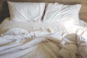 chaotisch bequeme weiße Bettwäsche und Kissenleben im Urlaub. Unordentliches, ungemachtes Bett mit weißem, zerknittertem Bettlaken und zwei unordentlichen Kissen in einem Hotelzimmer. feiertagswochenende sonntag hintergrund zur entspannung foto