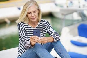 ältere Frau, die ihr Smartphone benutzt, sitzt in einem Seehafen. foto