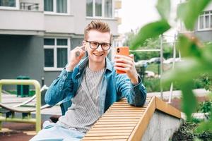 Portrait Mann in Hemd und Brille mit Kommunikation auf einem Smartphone, das auf einer Bank vor einem Hintergrund von Häusern sitzt foto