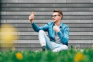 Mann mit Brille, der auf grünem Gras sitzt und auf einem grauen Wandhintergrund auf das Smartphone schaut foto