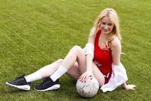 Die Blondine hält den Ball auf dem Fußballplatz zwischen ihren Beinen. foto