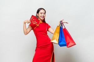 asiatische frau im traditionellen qipao-kleid, das taschen und rote umschläge im isolierten hellgrauen studiohintergrund für das chinesische einkaufskonzept des neuen jahres hält, text bedeutet viel glück, großer gewinn foto