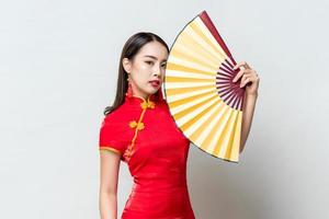 porträt der schönen asiatischen frau, die traditionelles rotes qipao-kleid trägt, das goldenen fan im isolierten hellgrauen hintergrund des studios für chinesisches neujahrskonzept hält foto