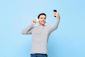 porträt eines glücklichen jungen gutaussehenden kaukasischen mannes mit kopfhörern, der musik hört, während er selfie auf isoliertem studioblauem hintergrund macht foto