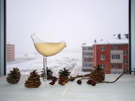 Hafermilch in einem dekorativen Glas in Form eines Vogels auf einem weißen Tisch mit Zapfen und Hagebutten. Schnee im Hintergrund foto