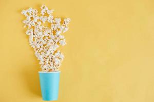 Popcorn in einem blauen Pappbecher auf gelbem Hintergrund verstreut. minimalistisches Konzept. Ansicht von oben. kopieren, leerer platz für text foto