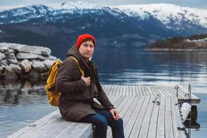 Mann mit einem gelben Rucksack, der einen roten Hut trägt, der auf einem Holzsteg auf dem Hintergrund von Berg und See sitzt foto