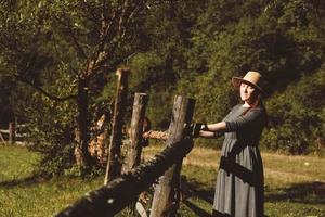 Frau mit Sommerkleid und Strohhut, die bei Sonnenuntergang in der Nähe eines Holzzauns auf einem Hintergrund aus Wald und Bäumen steht, mit geschlossenen Augen foto