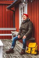 Reisender mit gelbem Rucksack sitzt in der Nähe des rot gefärbten Holzhauses foto