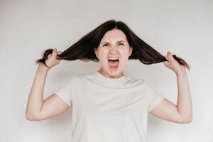 schockierte panische brünette frau in lässigem weißem t-shirt zieht ihre hände an ihren haaren und schreit verzweifelt und frustration, ihre augen voller entsetzen, offener mund. kopieren, leerer platz für text foto