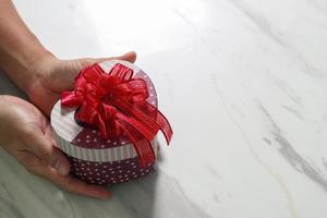 geschenk geben, man hand hält eine herzförmige geschenkbox in einer geste des gebens auf weißgrauem marmortischhintergrund foto