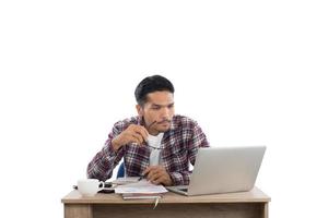 nachdenklicher junger Mann, der Laptop betrachtet, während er an seinem Arbeitsplatz lokalisiert auf weißem Hintergrund sitzt. foto