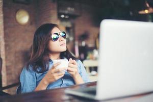 schöne junge hipster-frau, die in einem café sitzt und eine tasse kaffee hält, entspannt sich und spielt mit ihrem laptop, schaut weg, glücklich und macht spaß. Lifestyle-Konzept. foto