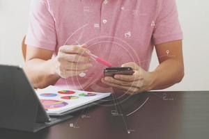 Geschäftsmann im rosafarbenen T-Shirt arbeitet mit Smartphone und digitalem Tablet-Computer auf Holzschreibtisch in modernem Büro mit virtuellem Symboldiagramm foto