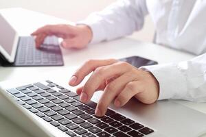 Nahaufnahme eines Geschäftsmannes, der ein digitales Tablet mit Tastatur und Laptop-Computer auf einem weißen Schreibtisch in einem modernen Büro eingibt foto