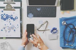 Draufsicht des intelligenten Arztes, der mit Handy und Laptop-Computer und Stethoskop auf dunklem Holzschreibtisch mit Symboldiagramm der virtuellen Realität arbeitet foto