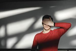 Junger Mann, der sein Gesicht mit der Hand in einem roten Pullover auf weißem Hintergrund mit Schatten aus dem Licht bedeckt. kopieren, leerer platz für text foto